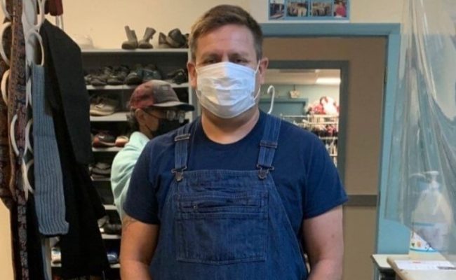 Guy in overalls and covid mask standing in front of clothing at Nourish.